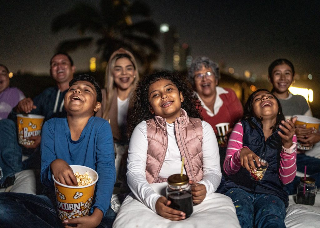 Family with adults and kids watching a movie outdoors on blankets and pillows laughing